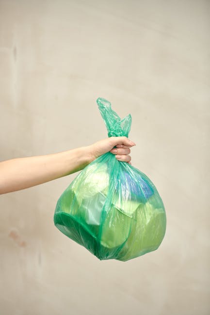 A person's arm extending from the left side of the image holding a green plastic rubbish bag by its knot, with the bag filled with miscellaneous waste and tied securely at the top. The bag appears slightly crumpled and semi-transparent, revealing some contents inside, with hints of blue and yellow visible through the plastic. The background is a plain, light-colored wall, creating a neutral setting that emphasizes the bag. This scene suggests an act of collection or disposal, typical of domestic or professional rubbish removal contexts, aligning with services provided by Waste Collection Bow for local authorities or private waste handling in the Bow area. The lighting is even and soft, without harsh shadows, highlighting the textured surface of the plastic bag and the smooth skin of the hand. This image visually relates to the process of binning waste, part of ongoing rubbish collection or on-site clearance efforts.