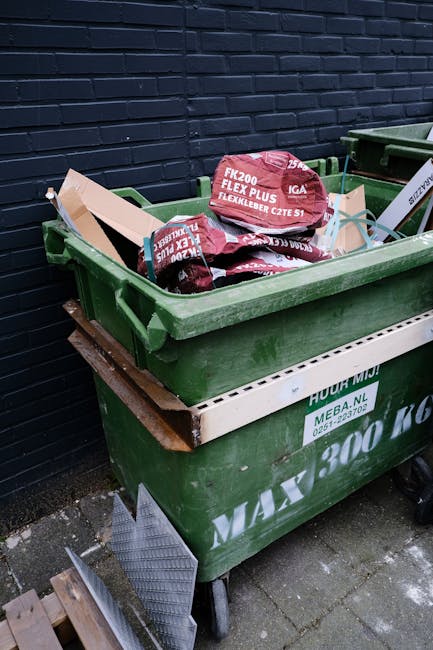 A large green waste bin positioned on a paved surface against a dark blue brick wall, filled with red and black packages of construction or garden materials labeled 'FK200 FLEXY PLUS' and 'GAZELLE.' The materials inside appear to be flexible piping or tubing. The bin has a white label with green text indicating it is part of a waste collection service, and it rests on a wheeled metal frame with a wooden plank acting as a support underneath. Adjacent to the bin, partially visible, is another green waste container. Natural lighting illuminates the scene, highlighting the textures of the plastic materials, the brick wall, and the paved ground, suggesting an outdoor setting associated with private waste disposal or rubbish removal services, such as those offered by Waste Collection Bow for the Devons Road estate in Bow.