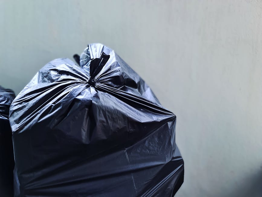 A person's arm extending from the left side of the image holding a green plastic rubbish bag by its knot, with the bag filled with miscellaneous waste and tied securely at the top. The bag appears slightly crumpled and semi-transparent, revealing some contents inside, with hints of blue and yellow visible through the plastic. The background is a plain, light-colored wall, creating a neutral setting that emphasizes the bag. This scene suggests an act of collection or disposal, typical of domestic or professional rubbish removal contexts, aligning with services provided by Waste Collection Bow for local authorities or private waste handling in the Bow area. The lighting is even and soft, without harsh shadows, highlighting the textured surface of the plastic bag and the smooth skin of the hand. This image visually relates to the process of binning waste, part of ongoing rubbish collection or on-site clearance efforts.